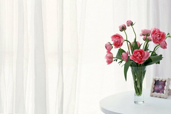 Peonies in a glass vase on a table in a room with white curtains