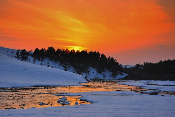 Winter Fluss vor dem Hintergrund von dunklen Bäumen und der erstaunlichen Schönheit eines hellen Sonnenuntergangs an einem wunderbaren Winterabend