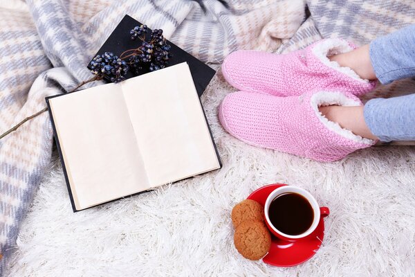 Legs of a girl in homemade pink ugg boots. A red cup with coffee on a red saucer. Oatmeal cookies. An open book. A warm plaid plaid and a white carpet with a large pile