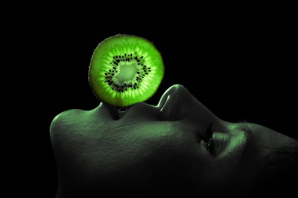 A man holds a slice of kiwi with his lips illuminated by a flashlight