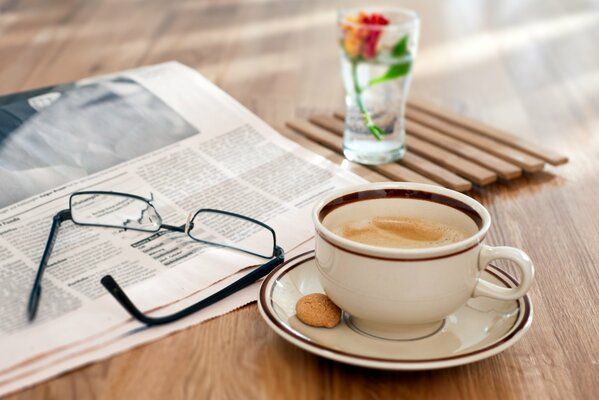 Una taza de café con una pequeña galleta está sobre la mesa, cerca se encuentran: periódico, vasos y un vaso con una flor