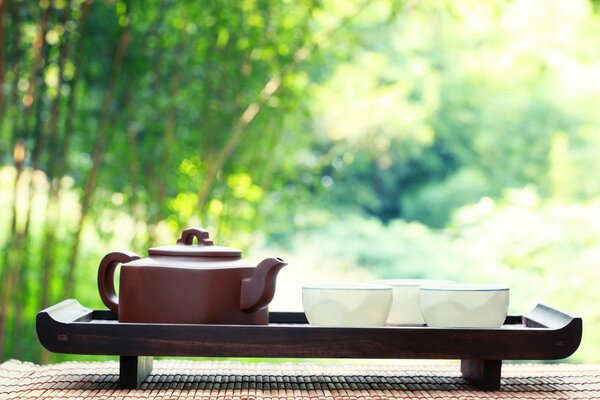 Una Tetera marrón y tres tazas blancas se colocan en una bandeja contra el fondo de la naturaleza