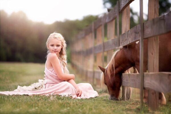 Drôle de fille assise sur l herbe, et derrière la clôture paissent un cheval