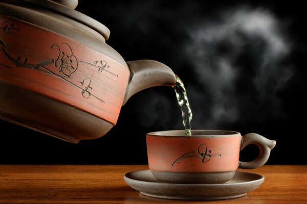 Grüner Tee aus der Teekanne, Dampf über der Tasse, Geschirr auf dem Tisch und schwarzer Nebel