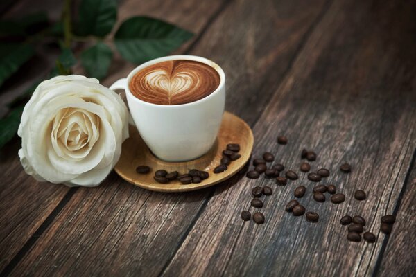 Weiße Rose auf einem Tisch mit einer weißen Tasse mit Schaum gefülltem Kaffee mit duftenden Kaffeebohnen daneben