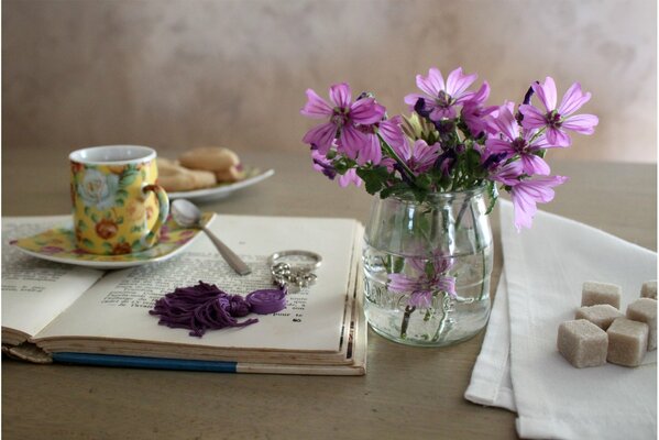 Tazza e piattino, biscotti e cubetti di zucchero. delicati fiori rosa in un vaso trasparente. libro aperto