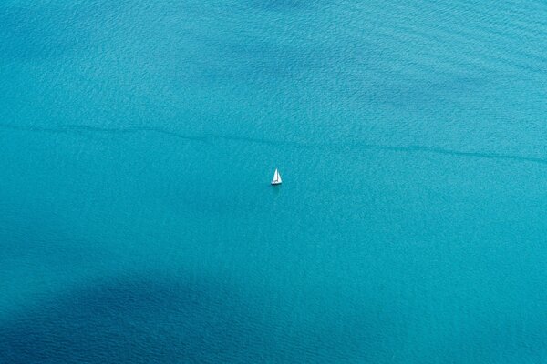 A boat is sailing far from the shore on the blue sea and only a white sail is visible