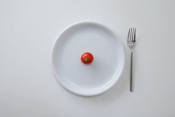 Sur une table blanche, une assiette blanche avec une tomate cerise et une fourchette