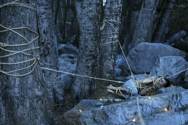 The guy is tied to trees on rocks in the forest