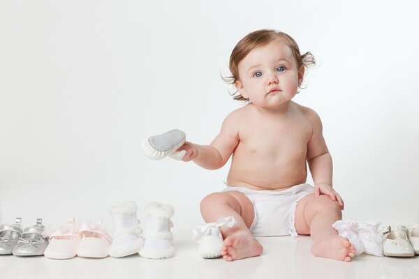 A small child is sitting on the floor in a diaper next to small shoe pairs of shoes