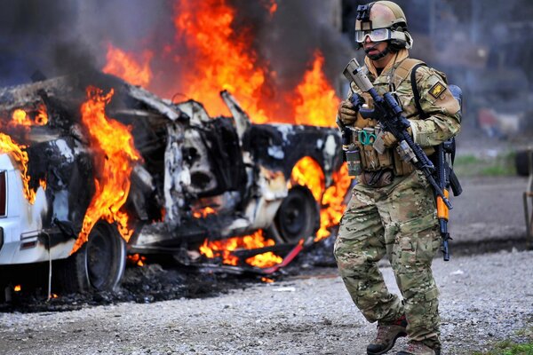 A soldier in full ammunition on the background of a burning car