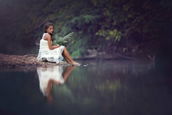 A sad girl is sitting on the shore with a bouquet of ferns
