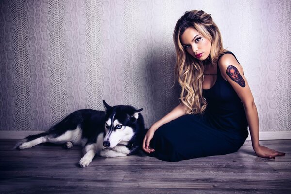 Una chica con un tatuaje se sienta con un perro Husky en un fondo de pared monocromático