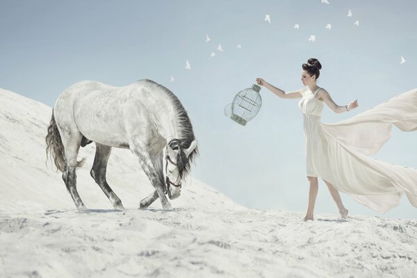 A woman in a white flying dress with a bird cage in her hands goes to a snow-white horse against the background of the Arctic desert