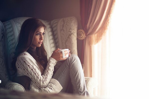 A girl in a chair with a mug on the background of the window