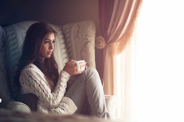 La jeune fille dans un fauteuil avec une tasse sur le fond de la fenêtre