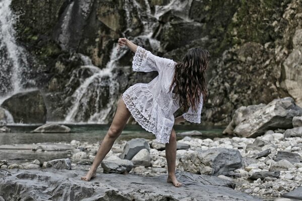 Una chica con un vestido blanco bailando en la orilla de un río de montaña