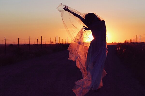 Esbelta chica con un vestido de gasa bailando en la carretera en el fondo de la puesta de sol de la noche