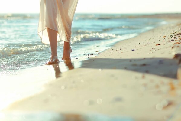 Nackte Füße eines am Strand entlang gehenden Mädchens in einem weißen Kleid