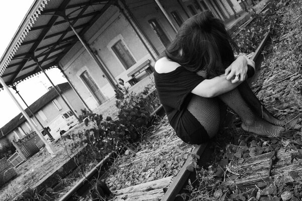 Ragazza triste in abito nero e collant si siede sui binari in una vecchia stazione ferroviaria in attesa di un treno