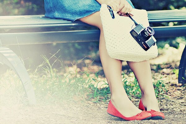 Legs in red ballet flats of a girl on a bench in a denim skirt with a camera and a knitted bag in her hands
