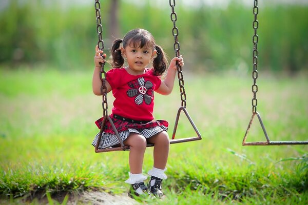 Una bambina dai capelli castani con le code di cavallo in un vestito rosso luminoso con un fiore si siede su un altalena tra il verde in una giornata di sole estiva