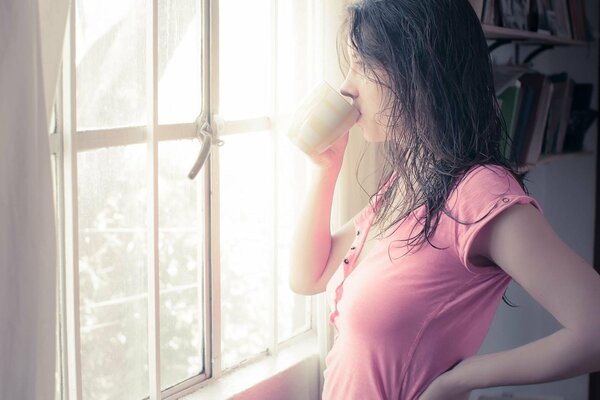 Brünette Mädchen mit nassen Haaren in einem rosa T-Shirt trinkt aus einem Becher am Fenster