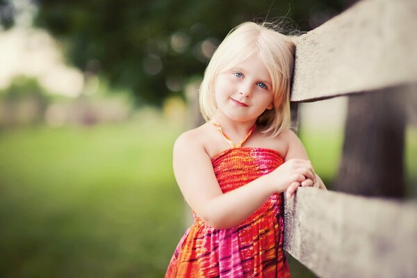 Una niña con un vestido de verano brillante y de buen humor está de pie en la cerca