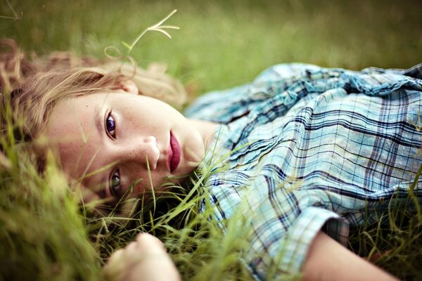 The blonde girl in the portrait is lying on the grass, thoughtfully looking at the camera