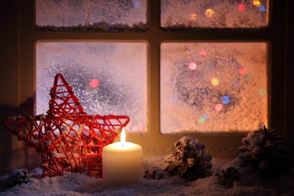 Estrella trenzada roja y vela ardiente con conos en el alféizar de la ventana de invierno