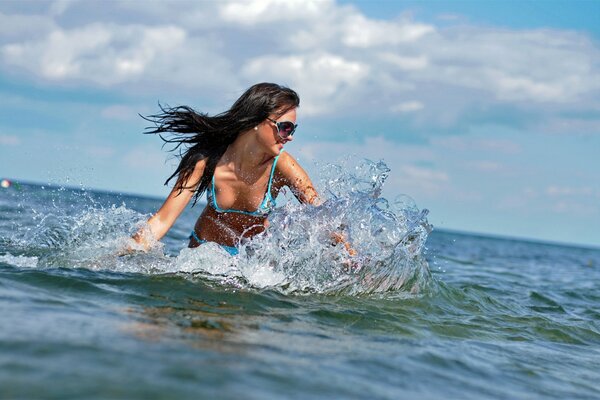Стройная девушка с темными волосами в очках , купается в море. Она улыбается, радуется хорошей солнечной погоде