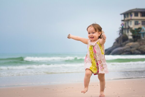 A happy little girl in a pink and yellow dress with flowers is walking along the wet coast and her hands are dirty in the sand