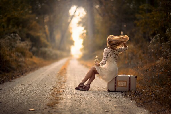 Ein Mädchen, das auf einem Koffer am Straßenrand sitzt. Der Wind flattert Ihr Haar