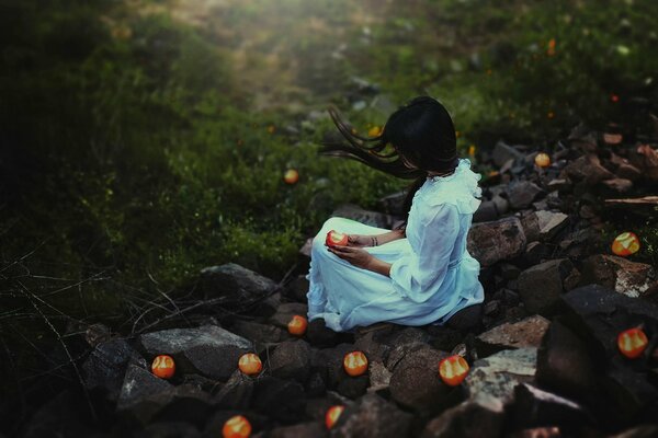 Una chica con un vestido blanco sentada en las piedras con una manzana en la mano