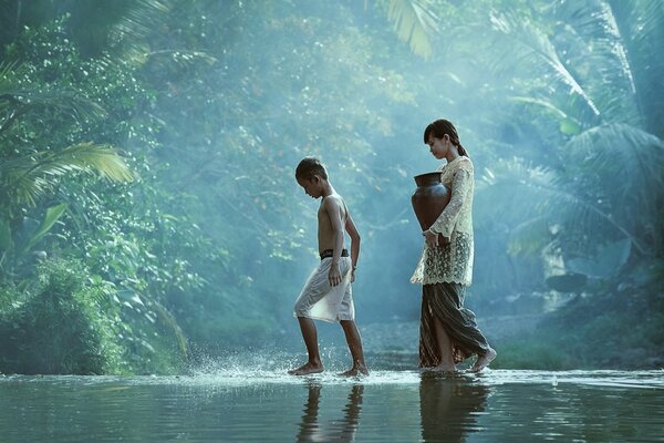 Niña con jarra y niño caminando por el río en la selva