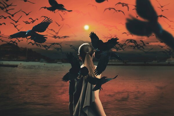 Against the background of the smooth water and the sunset sky, a girl in a dark dress is standing with her back to us, and crows are circling above her