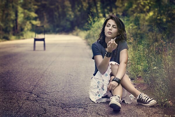 Девушка сидит на обочине дороги, на земле и курит, посреди дороги стоит стул
