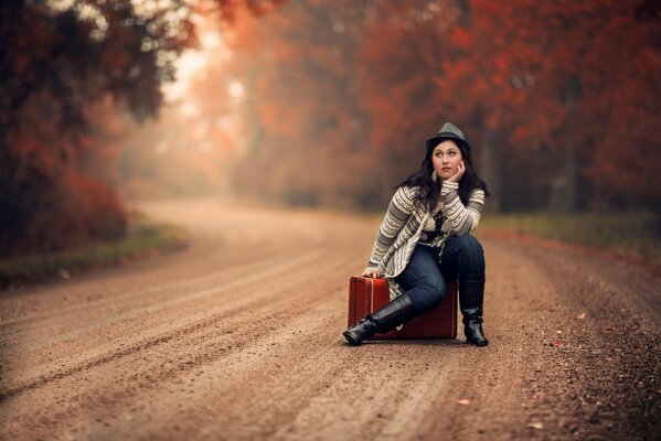 The girl is sitting on a suitcase on the road near the forest