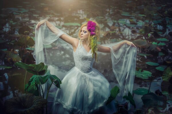 A girl in a white dress in a pond with water lilies with a bright flower in her hair