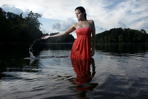 A brown-haired girl in a red sundress is standing knee-deep in water