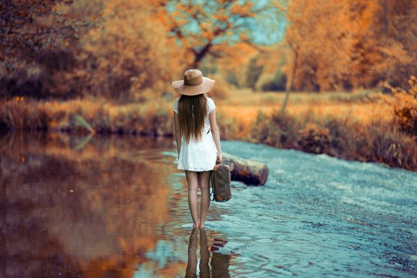 A girl in a white short dress and hat walks along the river with a suitcase, rear view