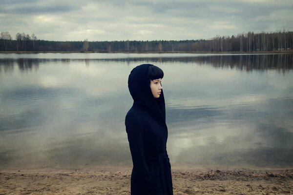 Dramatisches Foto eines Mädchens in einem schwarzen Mantel vor dem Hintergrund eines Sees und eines grauen Waldes ohne Laub