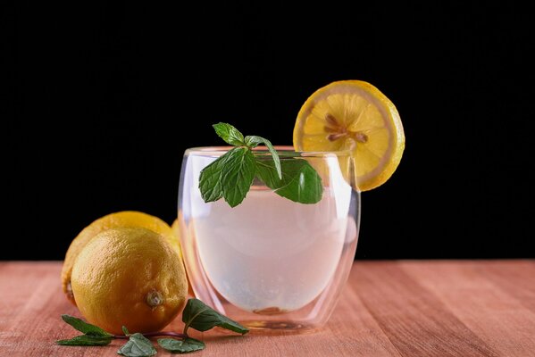 There is a glass with ice, a slice of lemon and mint on the table, lemons and mint sprigs are lying next to it