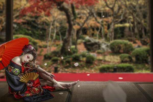 Ein Mädchen im asiatischen Outfit mit einem Regenschirm sitzt in einer Laube vor einem Hintergrund des Waldes