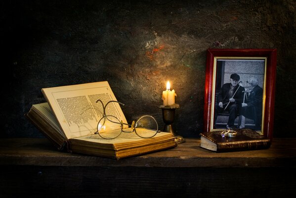 A book with glasses on the table near a candle and framed photos