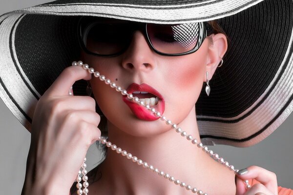 A girl with a hat and glasses bites a pearl necklace