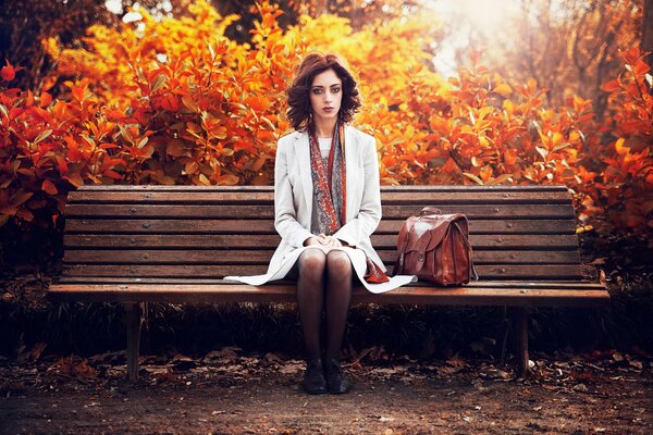 A brunette in a white raincoat sitting on a bench in an autumn park
