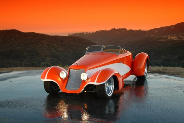 Orange Hot Rod steht auf nassem Asphalt vor dem Hintergrund von Hügeln und Sonnenuntergang