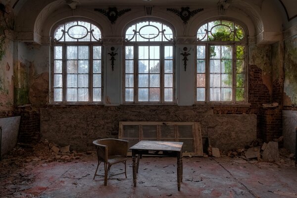 Mesa y sillón en una habitación con tres ventanas en un edificio abandonado