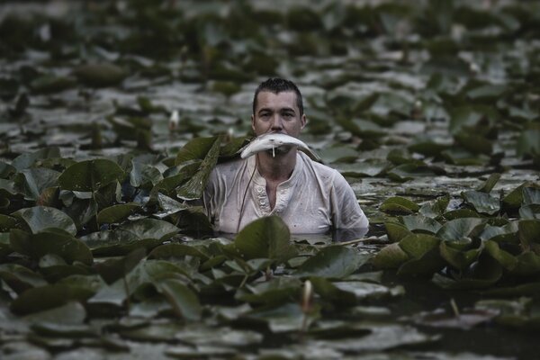 A guy with a fish in his mouth is standing in a pond